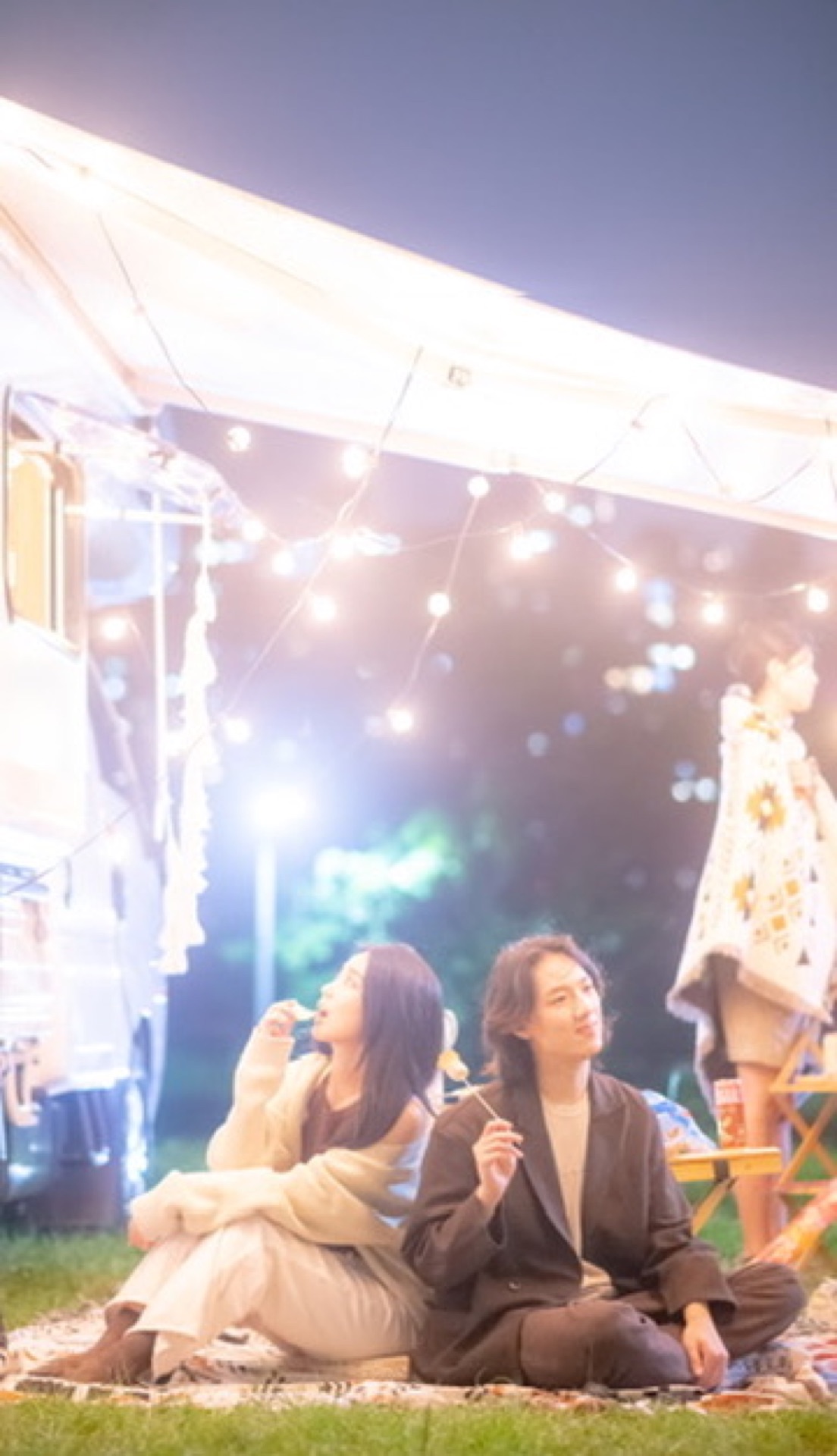 夜晚一對情侶背靠背坐在露營車天幕下的野餐墊上，溫暖球型燈串與鏡頭光暈，一旁為白色露營車，呈現情侶與好友揪團的夢幻露營車夜間氛圍