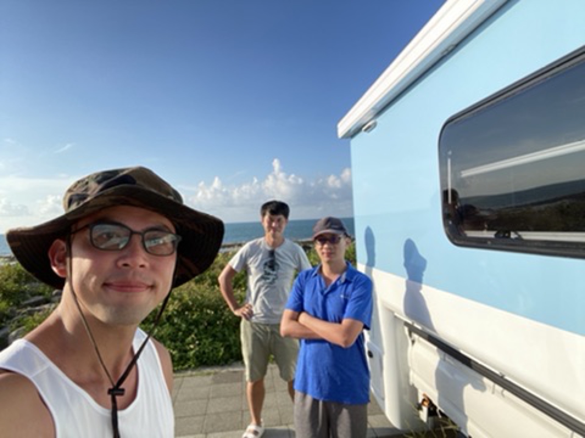 三名男性好友在墾丁海邊與露營車同框自拍，背景為沙灘與藍天，呈現陽光海線露營車出遊的青春回憶與愉悅體驗