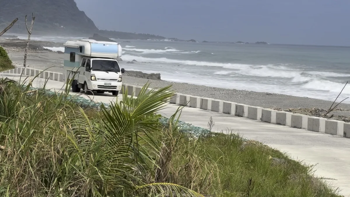 Joy Forest campervan driving slowly along a narrow coastal side road beside an open shoreline